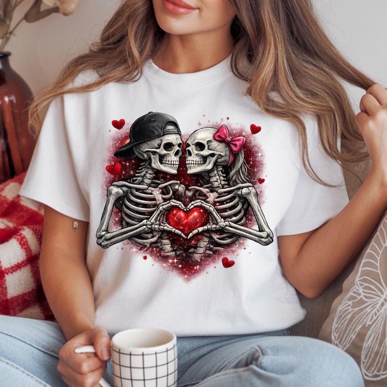 May include: White t-shirt featuring a graphic of two skeletons forming a heart with their hands. One skeleton wears a black cap, the other a pink bow. Red hearts and a red glitter background complete the design. A Valentine's Day themed tee.