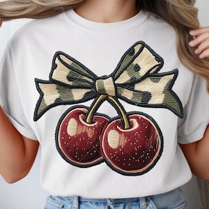 Può includere: T-shirt bianco con un disegno ricamato di due ciliegie e un fiocco. Le ciliegie sono di un rosso intenso con riflessi bianchi, e il fiocco ha un motivo mimetico nei toni del verde, beige e nero. Il disegno è centrato sulla maglietta.