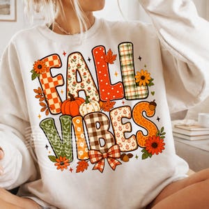 Puede incluir: Sudadera color crema con las palabras "FALL VIBES" en un diseño colorido y estampado. Las letras están decoradas con elementos temáticos de otoño como calabazas, hojas, flores y patrones de cuadros. Un gráfico con temática otoñal.