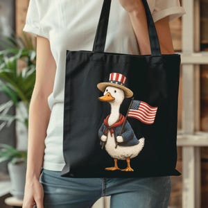 May include: Black tote bag featuring a cartoon duck dressed in patriotic attire. The duck wears a blue jacket, red scarf, and a hat with red and white stripes. It holds an American flag. The bag has black handles.