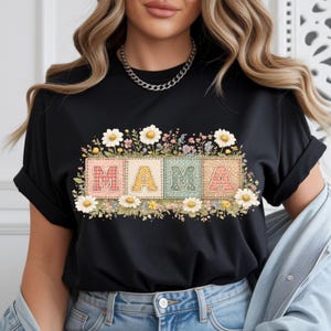 Peut inclure: T-shirt noir avec le mot "MAMA" dans un motif floral. Les lettres sont de couleurs pastel et entourées de marguerites et d'autres fleurs. Le t-shirt a des manches retroussées et un col rond.