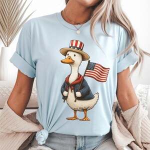 May include: Light blue t-shirt featuring a cartoon duck dressed in patriotic attire. The duck wears a top hat, a red scarf, and a blue jacket, and holds a small American flag. The design is a whimsical celebration of American pride.