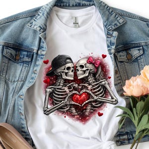 May include: White t-shirt featuring a graphic of two skeletons forming a heart with their hands. One skeleton wears a black cap, the other a pink bow. Red hearts and glittery accents surround the couple, perfect for Valentine's Day.