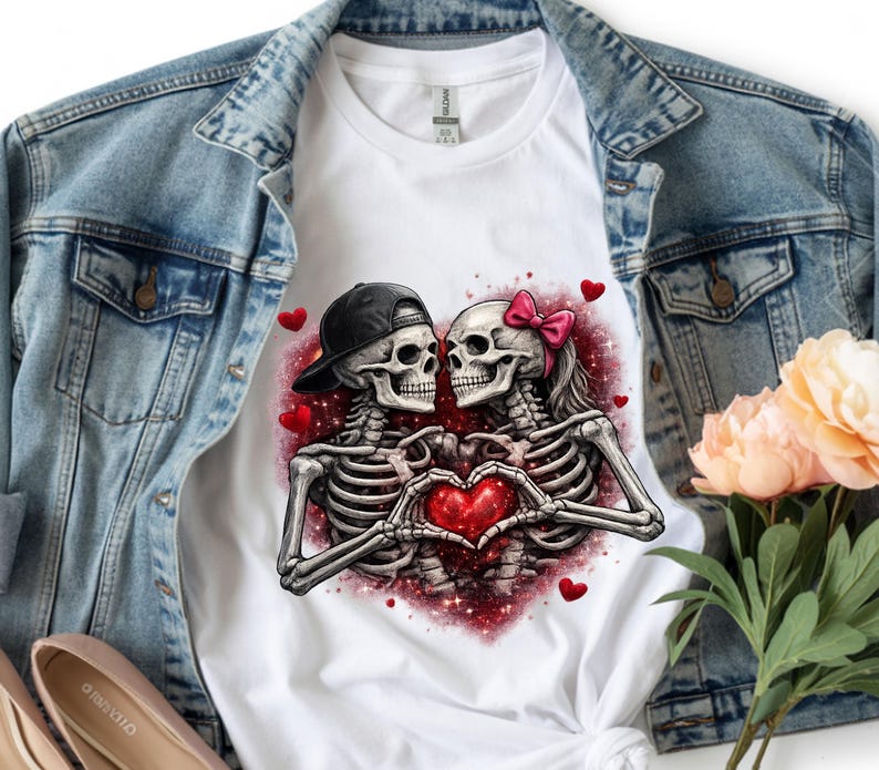 May include: White t-shirt featuring a graphic of two skeletons forming a heart with their hands. One skeleton wears a black cap, the other a pink bow. Red hearts and glittery accents surround the couple, perfect for Valentine's Day.