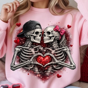 May include: Pink sweatshirt featuring a graphic of two skeletons in love, forming a heart with their hands. One skeleton wears a black baseball cap, the other a pink bow. Red hearts and glittery accents complete the design.