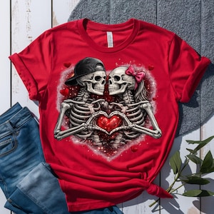 May include: Red t-shirt featuring a graphic of two skeletons forming a heart with their hands. One skeleton wears a black cap, the other a pink bow. The design is set against a heart-shaped background with red accents.