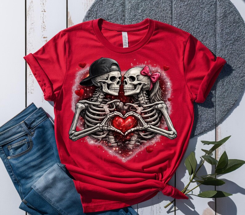 May include: Red t-shirt featuring a graphic of two skeletons forming a heart with their hands. One skeleton wears a black cap, the other a pink bow. The design is set against a heart-shaped background with red accents.