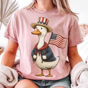 May include: Pink t-shirt featuring a cartoon duck dressed in patriotic attire. The duck wears a top hat with red and white stripes, a blue jacket, and a red scarf, holding an American flag. The design is a whimsical celebration of American pride.