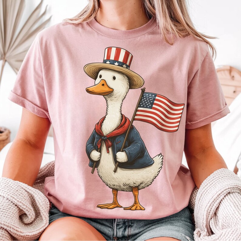 May include: Pink t-shirt featuring a cartoon duck dressed in patriotic attire. The duck wears a top hat with red and white stripes, a blue jacket, and a red scarf, holding an American flag. The design is a whimsical celebration of American pride.
