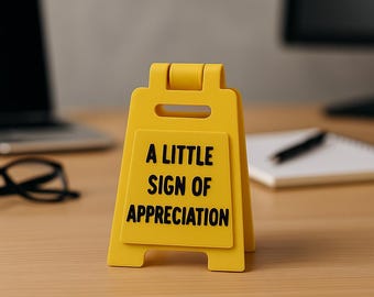 May include: A yellow plastic sign with the text "A LITTLE SIGN OF APPRECIATION" in black. The sign is in a triangular shape and is standing on a wooden surface. A laptop and glasses are in the background.