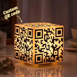 Pode incluir: Lâmpada em forma de cubo com design de código QR personalizado. A lâmpada emite uma luz amarela quente, destacando o padrão de código QR em preto e branco em todos os lados. O texto acima da lâmpada diz "Custom QR code".