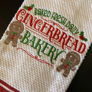 May include: White kitchen towel with red and green embroidered text that reads "Baked Fresh Daily Gingerbread Bakery" with two gingerbread men on either side of the text.