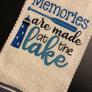 May include: White kitchen towel with blue and black stripes at the bottom. The towel has blue embroidery that says "Memories are made at the lake" with a lighthouse and a blue oar.