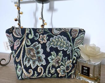 Op de afbeelding: Een zwarte en groene, met bloemen geprinte, rits-tas met de tekst "Clarofella" aan de zijkant. De tas ligt op een houten oppervlak naast een doorzichtige glazen fles met een witte bloem erop.