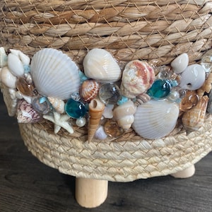 Op de afbeelding: Een ronde mand van gevlochten zeegras met een decoratieve band van diverse schelpen, kleine witte zeesterren en doorschijnende blauwe en heldere glaskralen. De mand rust op drie korte, lichtgekleurde cilindervormige houten pootjes.