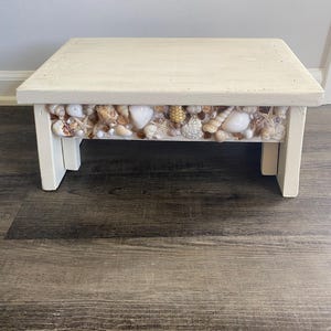 Op de afbeelding: Een kleine, vierkante, crèmekleurige houten kruk. De voorkant van de kruk is versierd met een verscheidenheid aan schelpen. De kruk heeft vier poten en is geschikt voor decoratief of functioneel gebruik.
