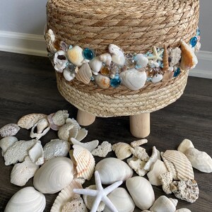 Op de afbeelding: Een ronde, geweven kruk met een decoratieve band van schelpen, glazen kralen en kleine zeesterren. De kruk heeft drie houten poten en is omgeven door een verzameling verschillende schelpen en een witte zeester.