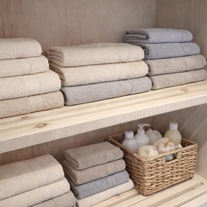 May include: A wooden cabinet with shelves stacked with folded bath towels in neutral colours. A woven basket on the lower shelf contains toiletries. The towels are in shades of beige, grey, and white, creating a spa-like aesthetic.