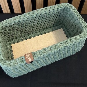May include: A rectangular sage green crocheted basket with a light-colored wooden base. The basket features a thick, textured knit pattern and a small brown leather-like tag on the side with the text "Lilea Handmade" printed in white.