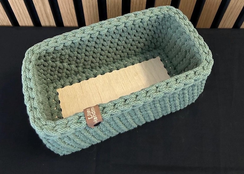 May include: A rectangular sage green crocheted basket with a light-colored wooden base. The basket features a thick, textured knit pattern and a small brown leather-like tag on the side with the text "Lilea Handmade" printed in white.