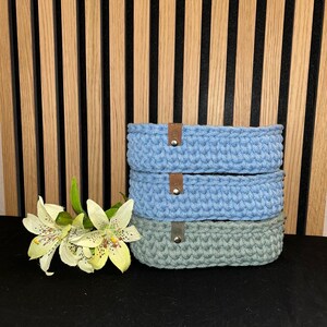 May include: A stack of three oval-shaped crocheted baskets. The top two baskets are light blue, and the bottom basket is sage green. Each basket features a small brown leather-like tab secured with a silver-colored metal rivet on the front side.