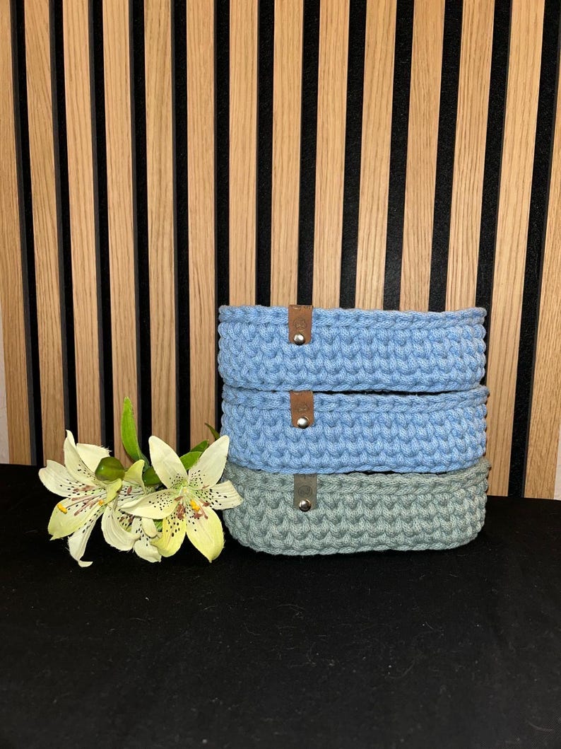 May include: A stack of three oval-shaped crocheted baskets. The top two baskets are light blue, and the bottom basket is sage green. Each basket features a small brown leather-like tab secured with a silver-colored metal rivet on the front side.