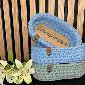 May include: Two oval crochet baskets stacked vertically, one light blue and one sage green, each featuring a small brown leather tag with a metal rivet. The top basket contains a light brown wooden base with the text Brillenparkplatz engraved.