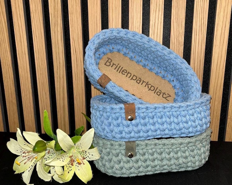 May include: Two oval crochet baskets stacked vertically, one light blue and one sage green, each featuring a small brown leather tag with a metal rivet. The top basket contains a light brown wooden base with the text Brillenparkplatz engraved.