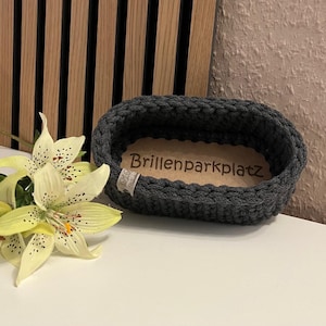 May include: An oval, dark gray crocheted basket with a light brown wooden base featuring the engraved text Brillenparkplatz. A small white fabric tag is attached to the side. Next to the basket is a pale yellow lily with dark speckles.