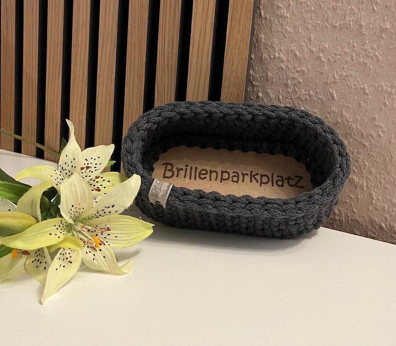 May include: An oval, dark gray crocheted basket with a light brown wooden base featuring the engraved text Brillenparkplatz. A small white fabric tag is attached to the side. Next to the basket is a pale yellow lily with dark speckles.