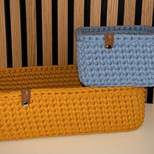 May include: Two rectangular storage baskets made of thick, chunky crochet yarn. One basket is mustard yellow and the other is light blue. Each basket features a small brown leather-like tab attached to the front rim with a silver metal rivet.