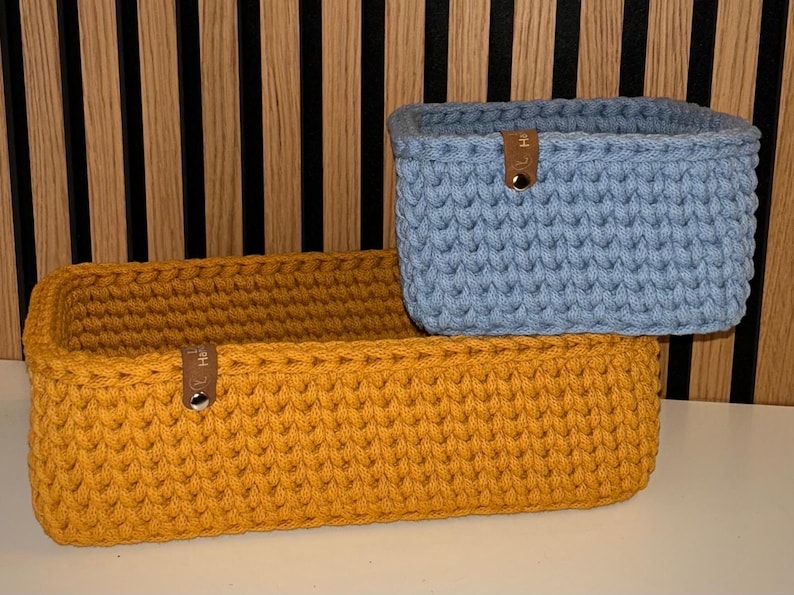 May include: Two rectangular storage baskets made of thick, chunky crochet yarn. One basket is mustard yellow and the other is light blue. Each basket features a small brown leather-like tab attached to the front rim with a silver metal rivet.