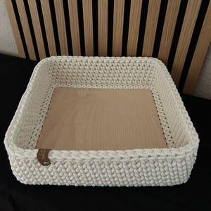 May include: A square storage basket featuring a thick, cream-colored crocheted yarn border and a flat, light-toned wooden base. A small, brown leather-like tag is attached to the side of the crocheted wall, adding a subtle contrast to the design.