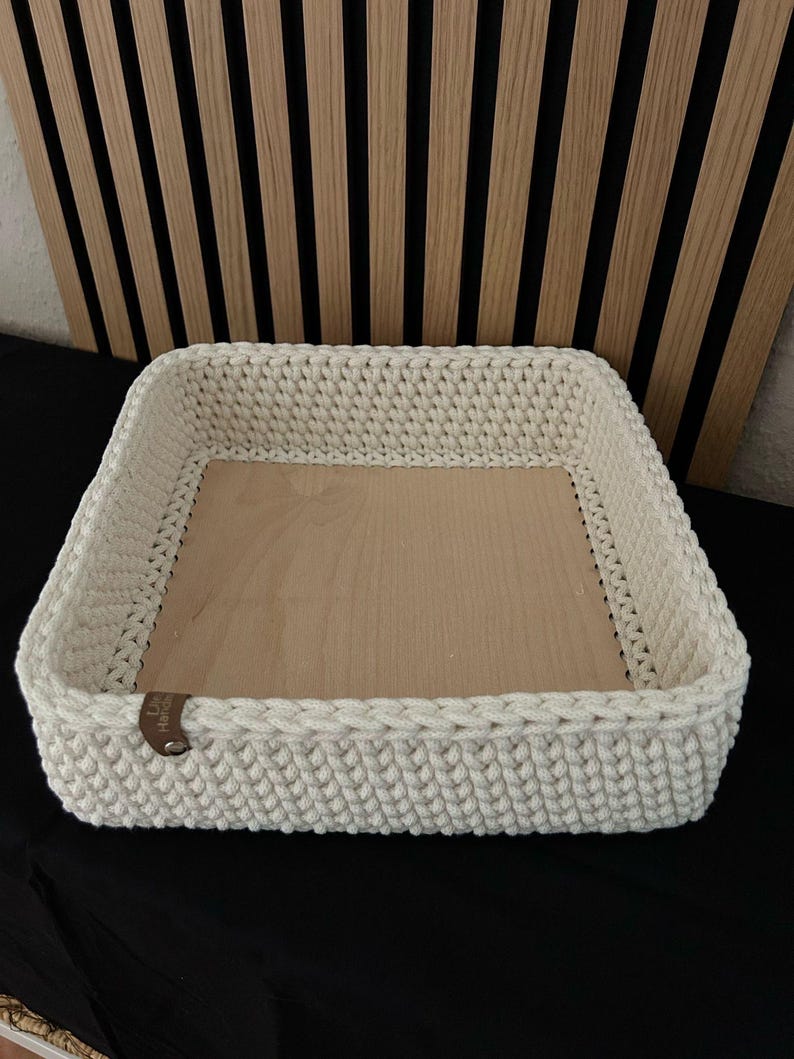 May include: A square storage basket featuring a thick, cream-colored crocheted yarn border and a flat, light-toned wooden base. A small, brown leather-like tag is attached to the side of the crocheted wall, adding a subtle contrast to the design.