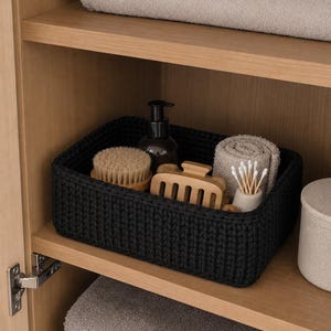 May include: A rectangular black crocheted storage basket containing a dark brown pump bottle, a round wooden brush, a light wooden hair claw clip, a small beige ceramic container with cotton swabs, and a rolled beige washcloth.
