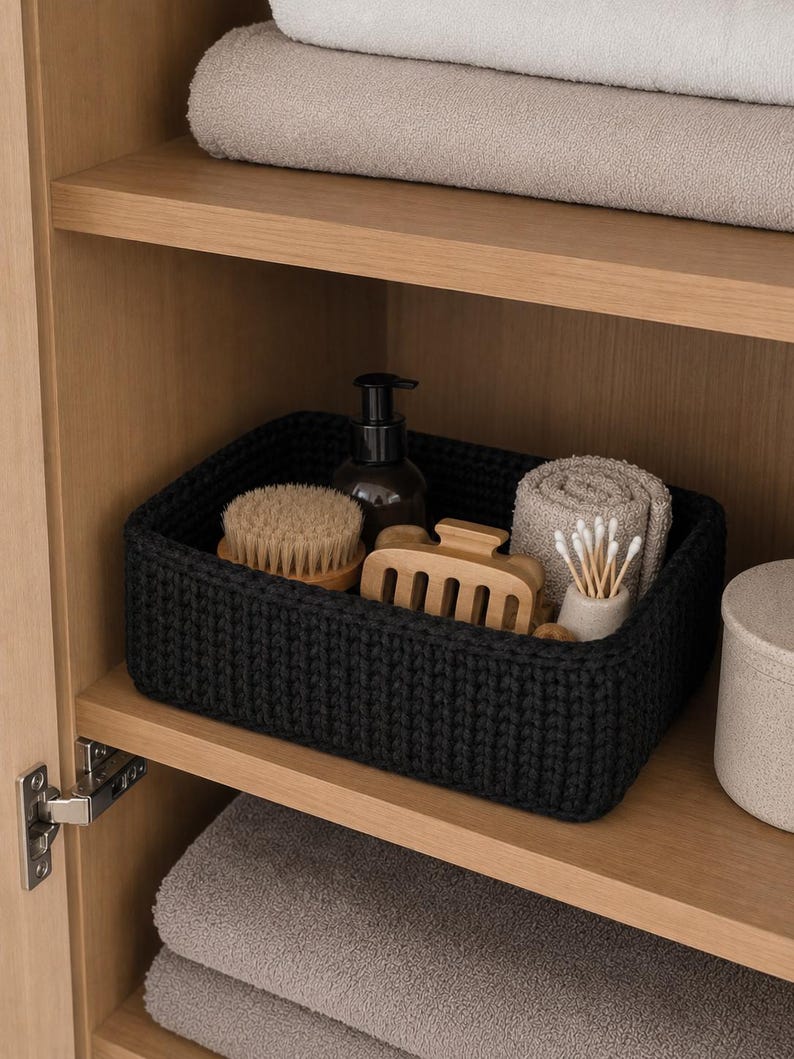 May include: A rectangular black crocheted storage basket containing a dark brown pump bottle, a round wooden brush, a light wooden hair claw clip, a small beige ceramic container with cotton swabs, and a rolled beige washcloth.