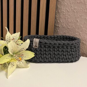 May include: An oval-shaped dark gray crocheted basket made of thick yarn. The basket features a small, light-colored rectangular tag attached to the side with a metal rivet, displaying a logo with the letter G and a small floral icon.