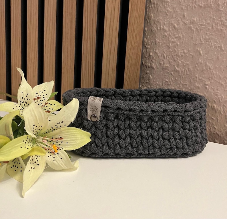 May include: An oval-shaped dark gray crocheted basket made of thick yarn. The basket features a small, light-colored rectangular tag attached to the side with a metal rivet, displaying a logo with the letter G and a small floral icon.