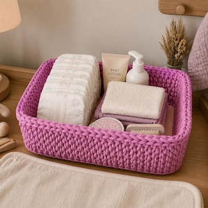 May include: A rectangular, bright purple, chunky knit storage basket containing a stack of disposable diapers, a tube of baby lotion, a white pump bottle, a folded beige washcloth, a small round container, and a wooden baby hairbrush.