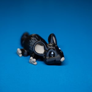 Peut inclure: Une petite figurine de souris noire brillante avec de grandes oreilles arrondies présentant des détails intérieurs beige clair. La souris a des accents blancs sur les yeux et le museau, avec de petites pattes claires visibles en position accroupie.