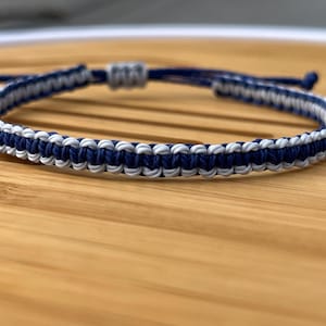 May include: A navy blue and silver braided bracelet. The bracelet is made with two different colors of cord and is tied with a knot at the end.