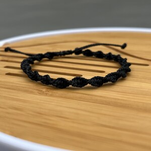May include: A black macrame bracelet with a series of knots forming a spiral pattern. The bracelet is made of a woven material and has adjustable ties on each end. The bracelet is displayed on a wooden surface.