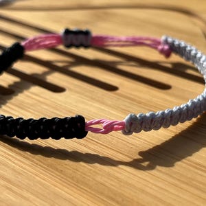 May include: A black, white, and pink braided friendship bracelet. The bracelet is made of three strands of cord, each a different color. The cords are braided together to create a unique and stylish design.