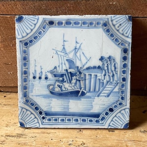 May include: A blue and white Delft tile depicting a sailing ship and figures on a dock. The tile has a decorative border with a blue and white pattern.
