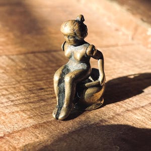 Pu&ograve; includere: Una piccola statuetta dorata di una donna seduta su una base rotonda. La statuetta &egrave; dettagliata e ha un aspetto vintage.