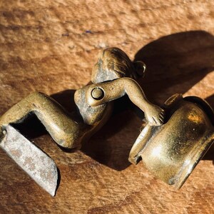 Pu&ograve; includere: Una statuetta in ottone di una persona seduta su una pentola. La statuetta &egrave; dettagliata e ha una finitura rustica.