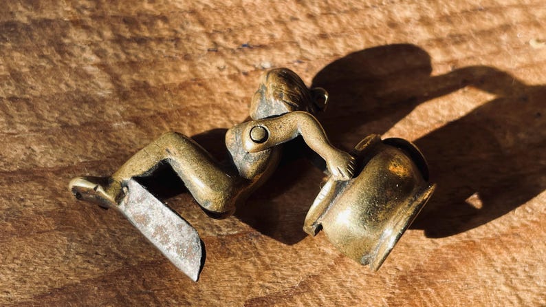 Pu&ograve; includere: Una statuetta in ottone di una persona seduta su una pentola. La statuetta &egrave; dettagliata e ha una finitura rustica.