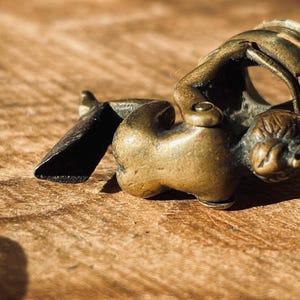Pu&ograve; includere: Una piccola figurina in metallo dorato che rappresenta una persona che tiene un oggetto curvo. La figurina &egrave; sdraiata su una superficie di legno.