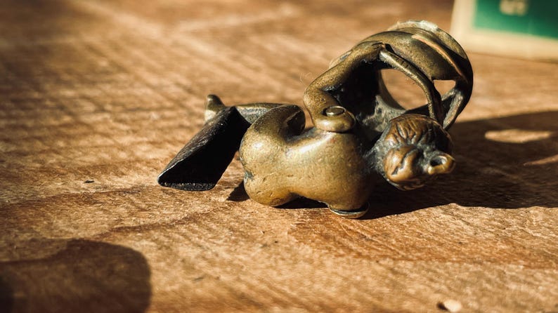 Pu&ograve; includere: Una piccola figurina in metallo dorato che rappresenta una persona che tiene un oggetto curvo. La figurina &egrave; sdraiata su una superficie di legno.