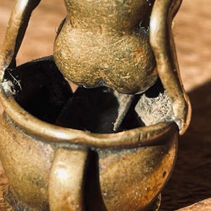 Pu&ograve; includere: Una piccola scultura in metallo dorato che rappresenta una persona seduta in una pentola. La scultura &egrave; dettagliata e ha un aspetto rustico e invecchiato.
